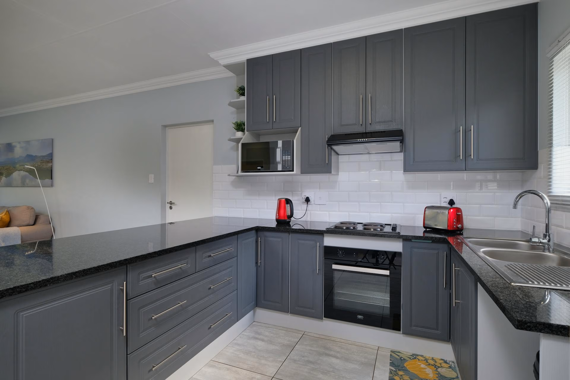 Modern kitchen with gray cabinets, black countertops, red kettle, toaster, and stainless steel sink.