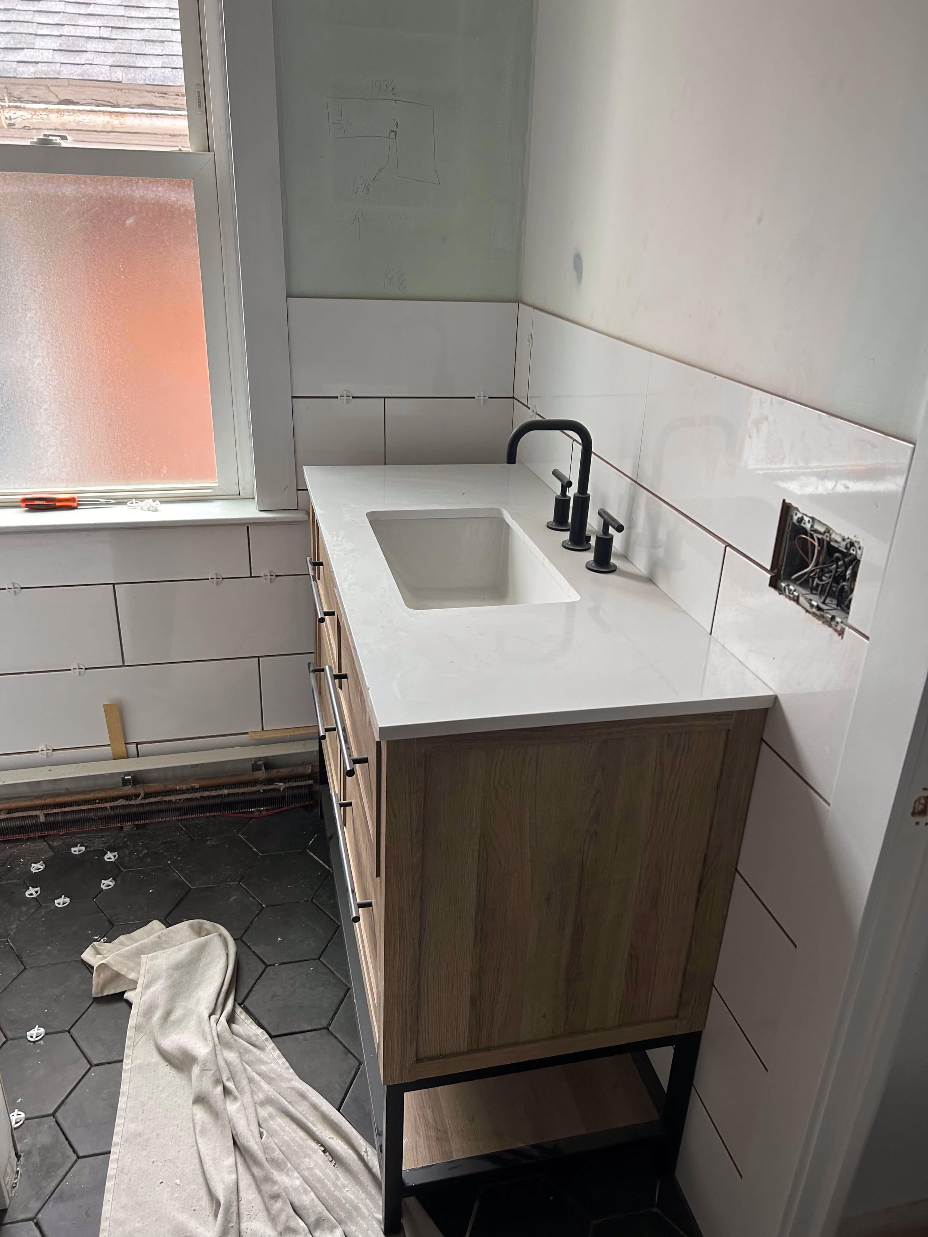 Wood vanity with white countertop and black faucet in partially tiled bathroom under renovation.