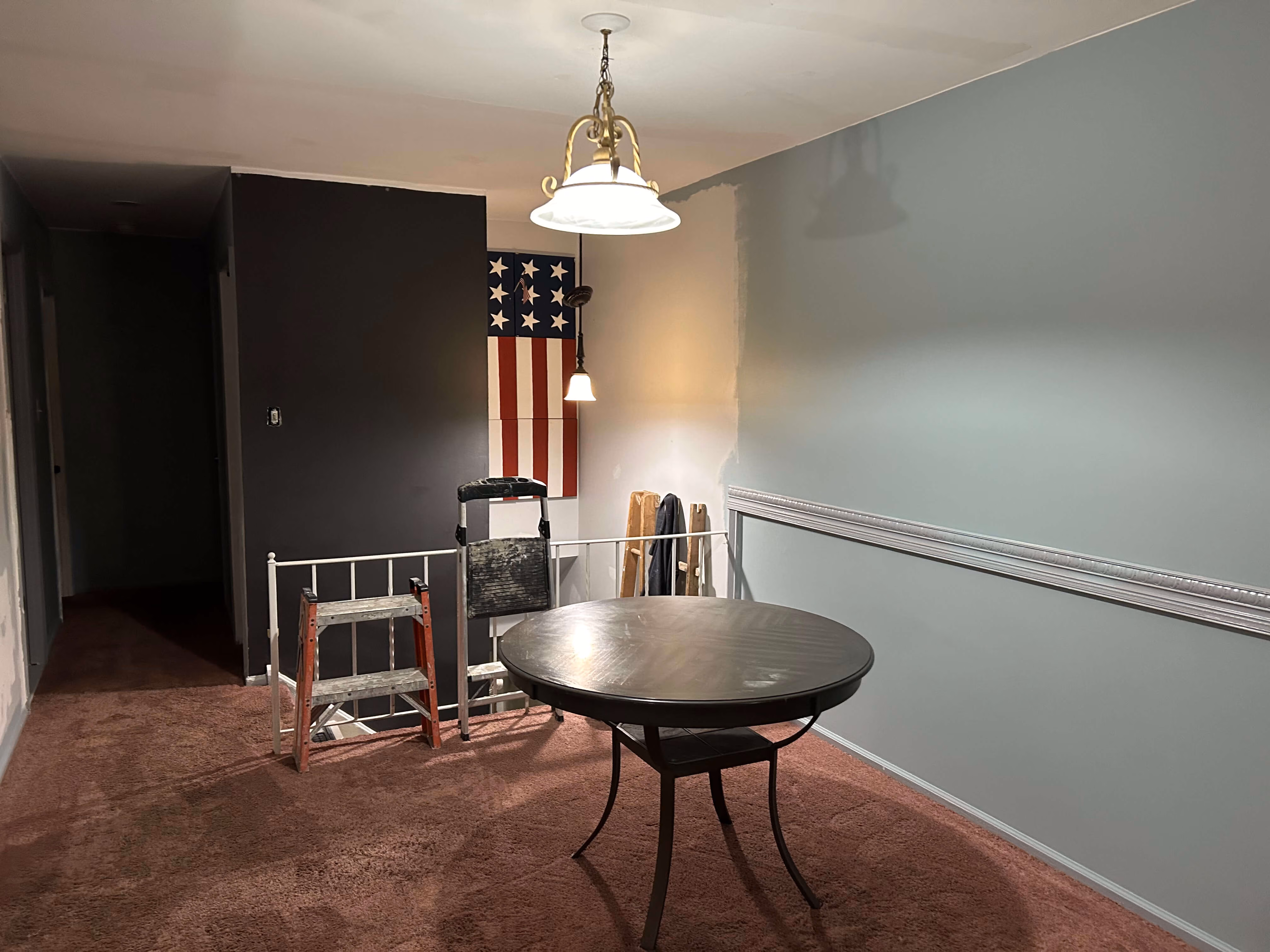 Room with brown carpet, round table, ladder, and partially painted walls with American flag artwork.