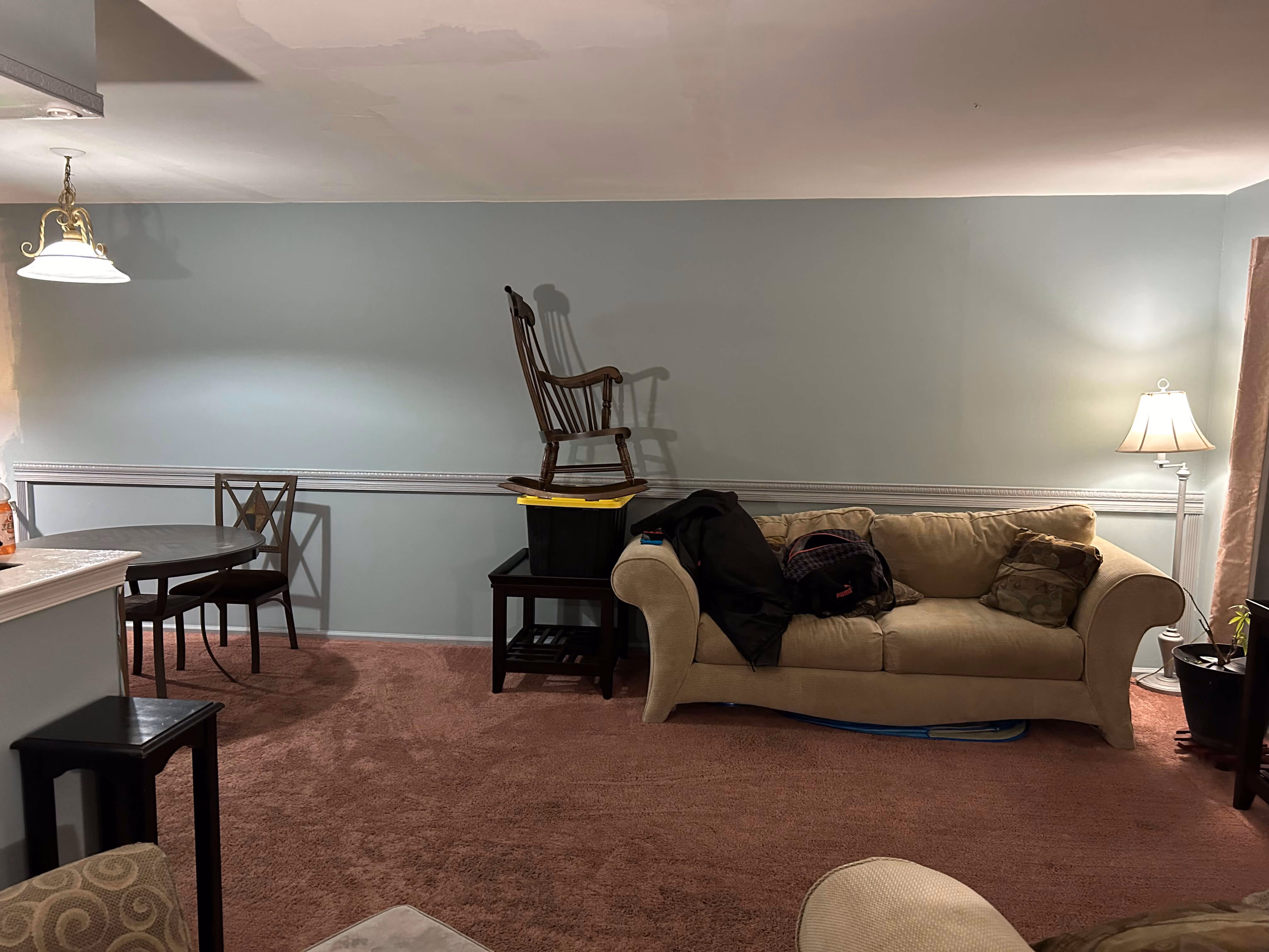 Living room with beige couch, side table holding plastic bin and rocking chair, dining table with chairs.