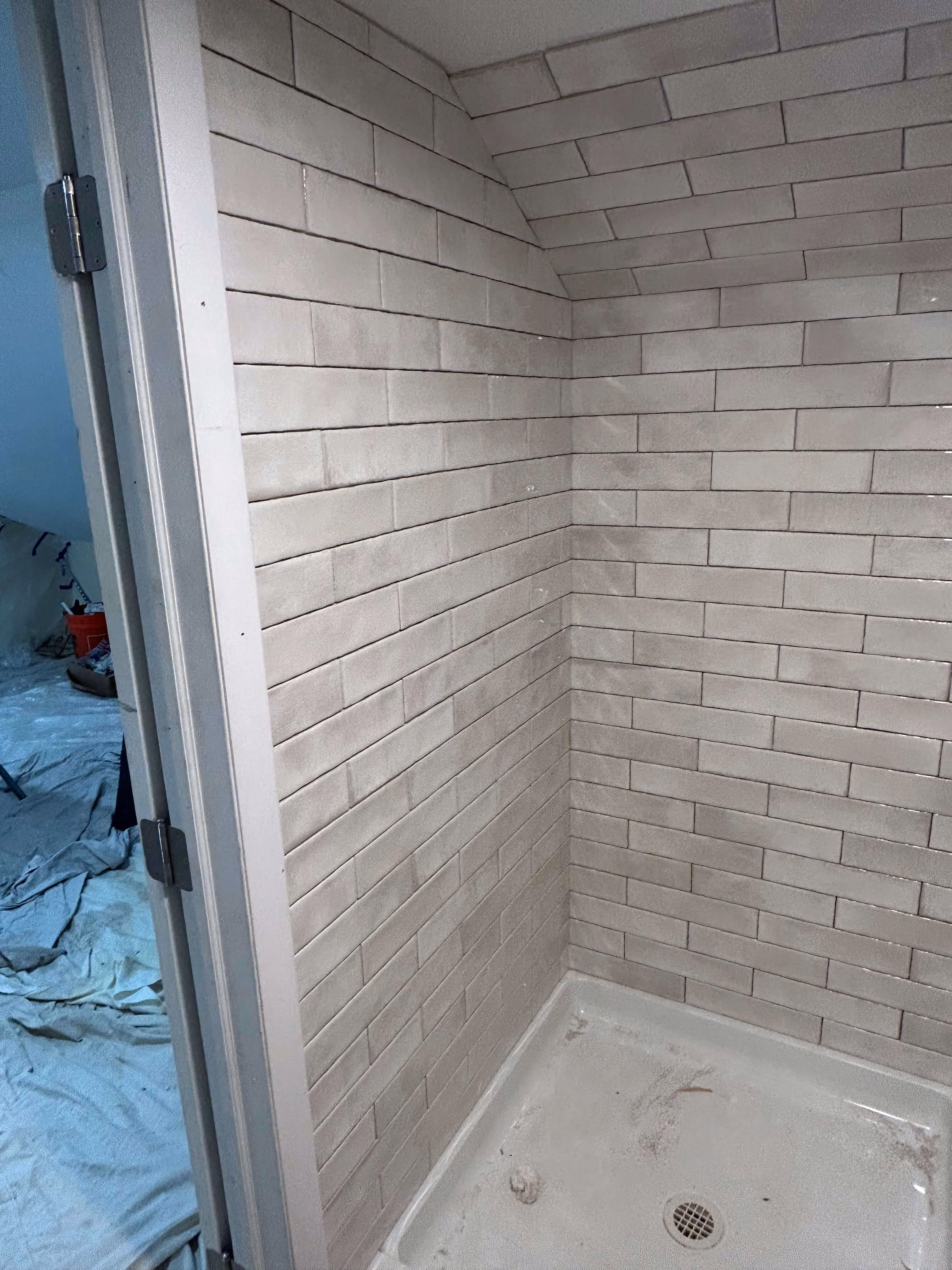 Corner of a tiled shower with light gray rectangular tiles and a white shower base with a drain.