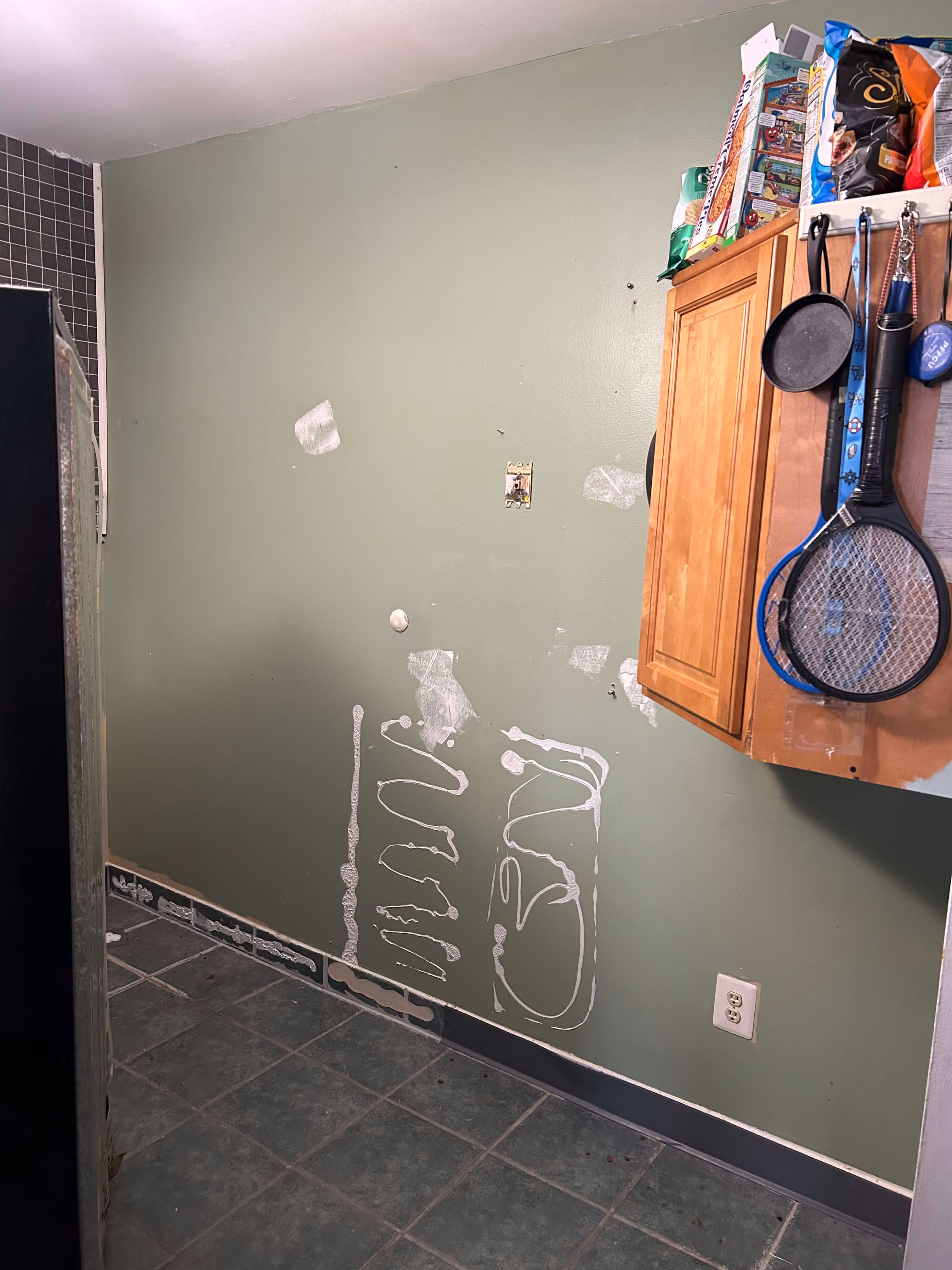 Green wall with white patch repairs, an empty socket, and a wooden cabinet with hanging items in a tiled room.