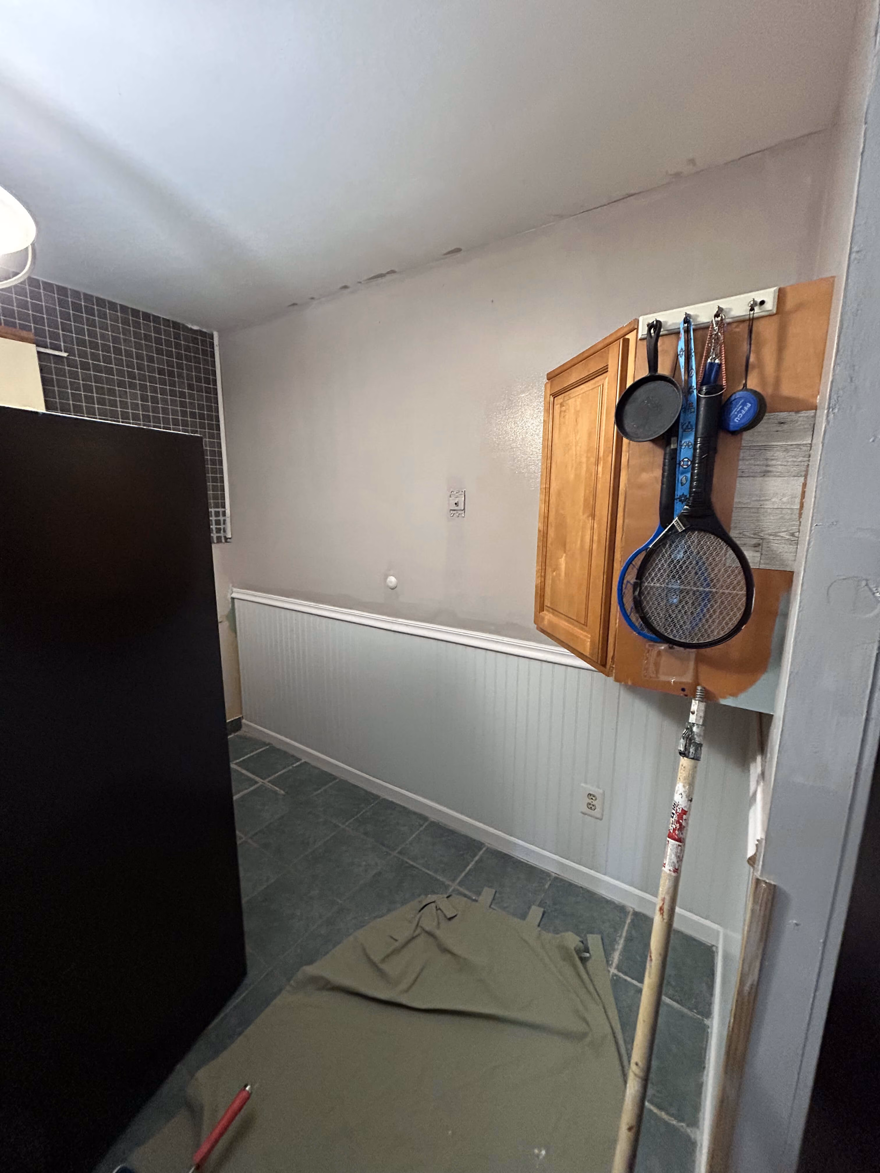 Corner of a kitchen with black fridge, hanging utensils, wooden cabinet, tiled floor, and draped cloth on floor.