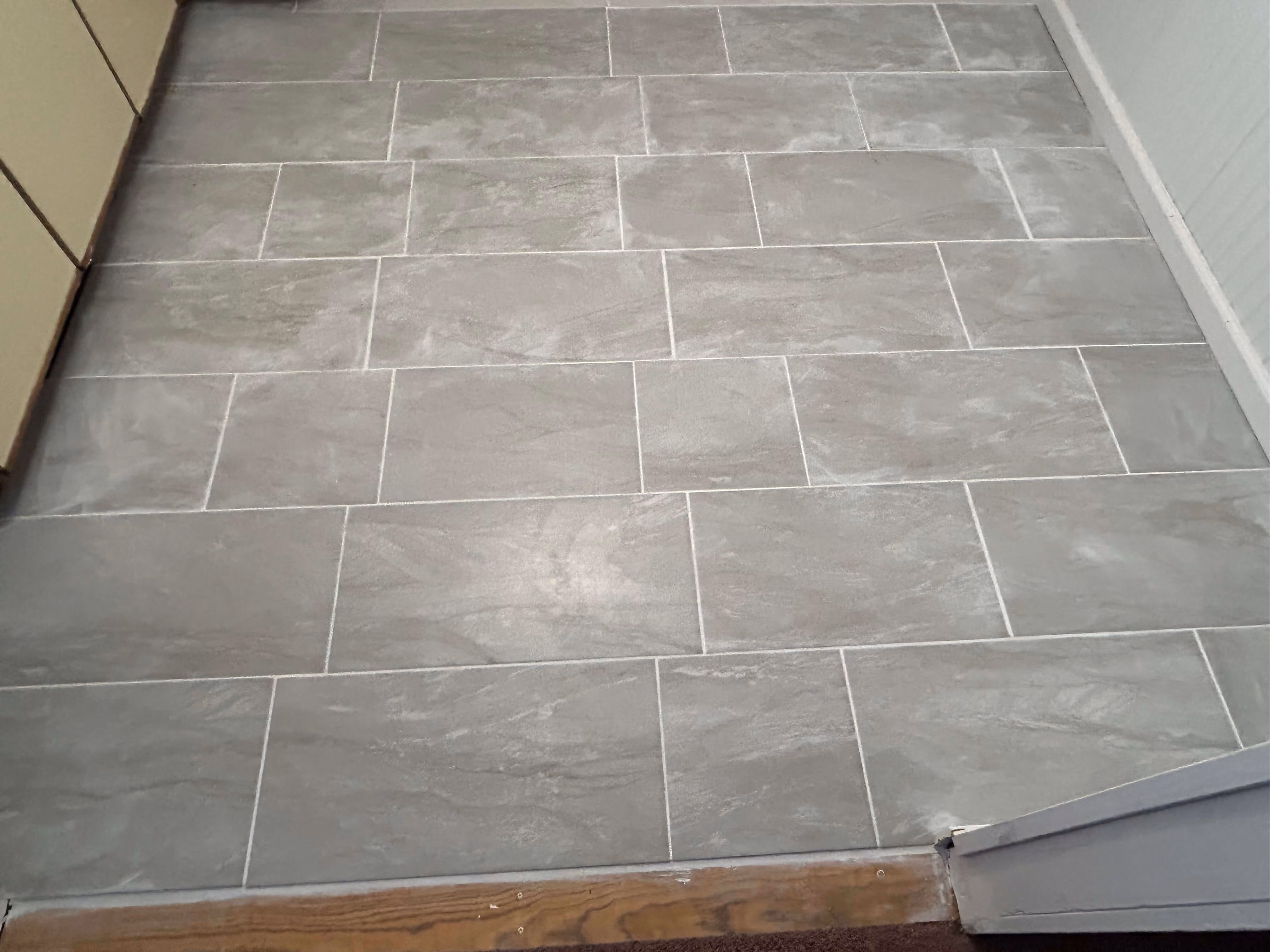 Floor with rectangular gray ceramic tiles arranged in a staggered pattern next to wood and carpet.
