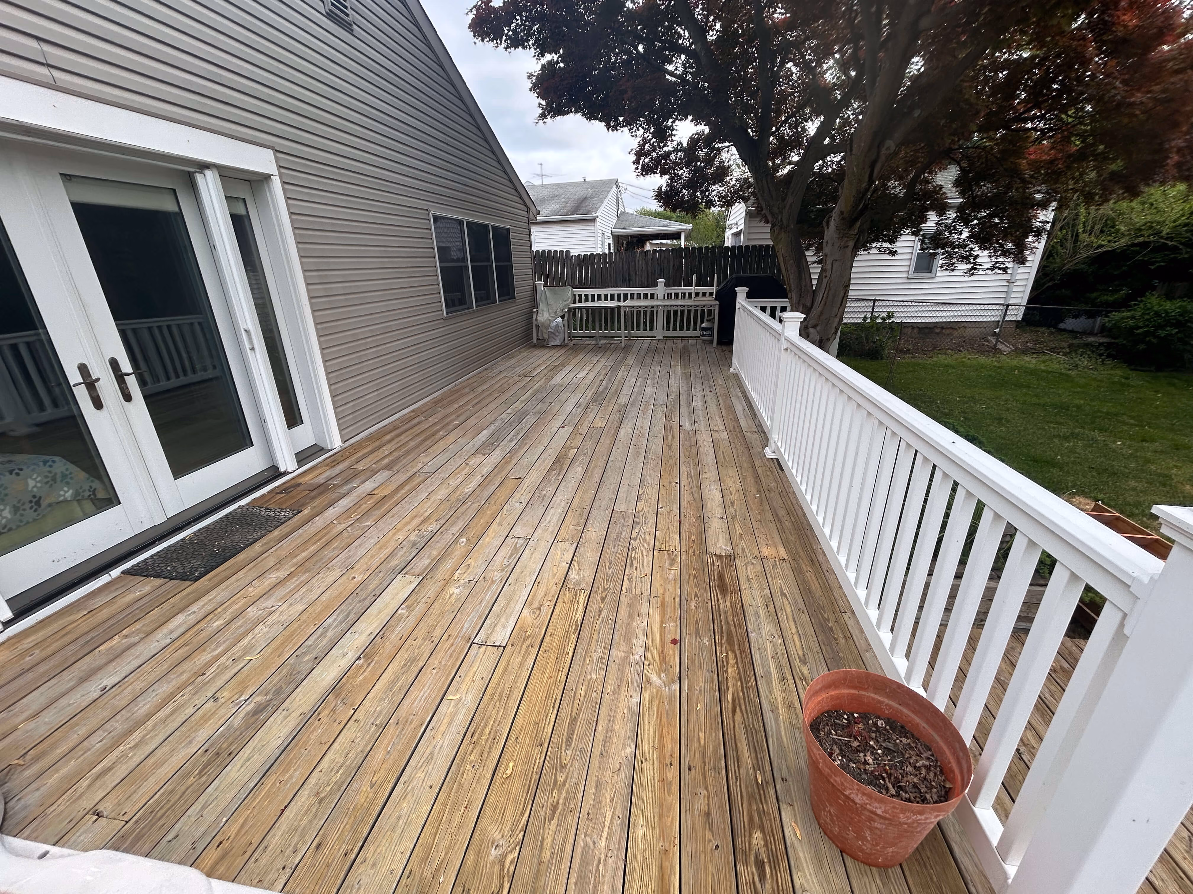 Wooden deck with white railing, potted plant, tree, and house with glass doors in backyard.