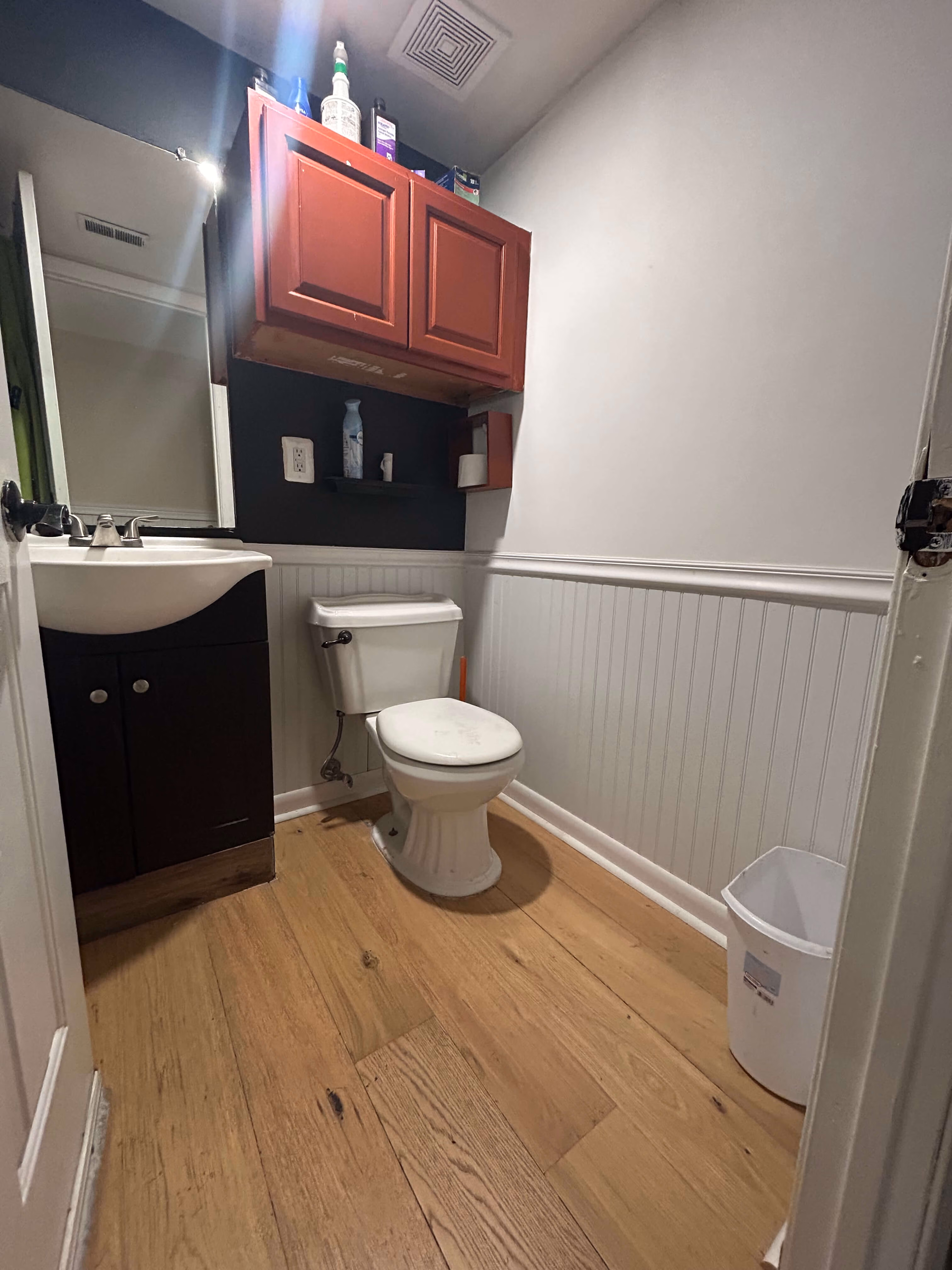 Small bathroom with wooden floor, white toilet, dark sink cabinet, red wall cabinet, and white trash bin.