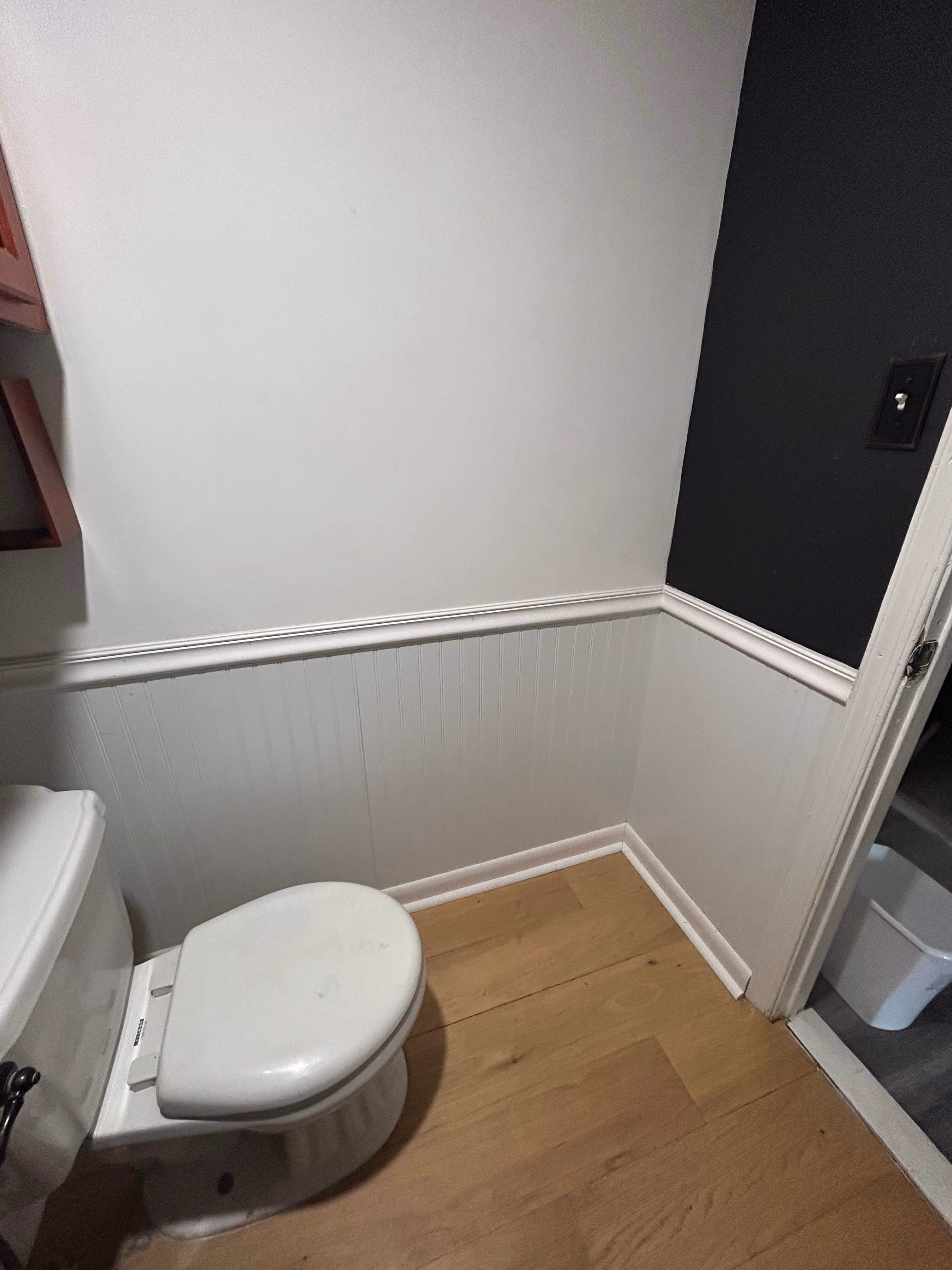 Small bathroom corner with a white toilet, light wood floor, white paneling, and part black wall.