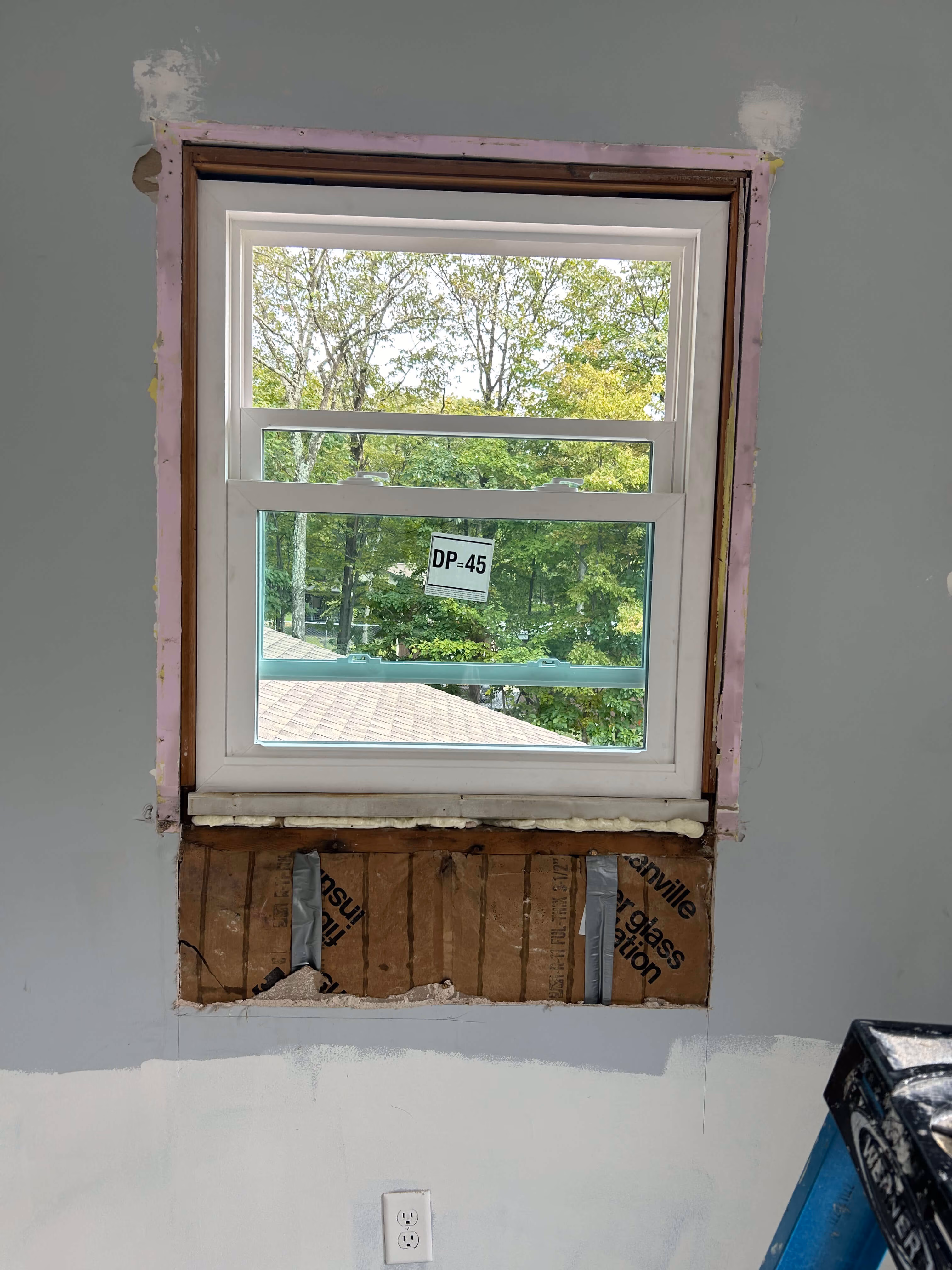 Half-installed window with exposed insulation and wall under construction, green trees visible outside.