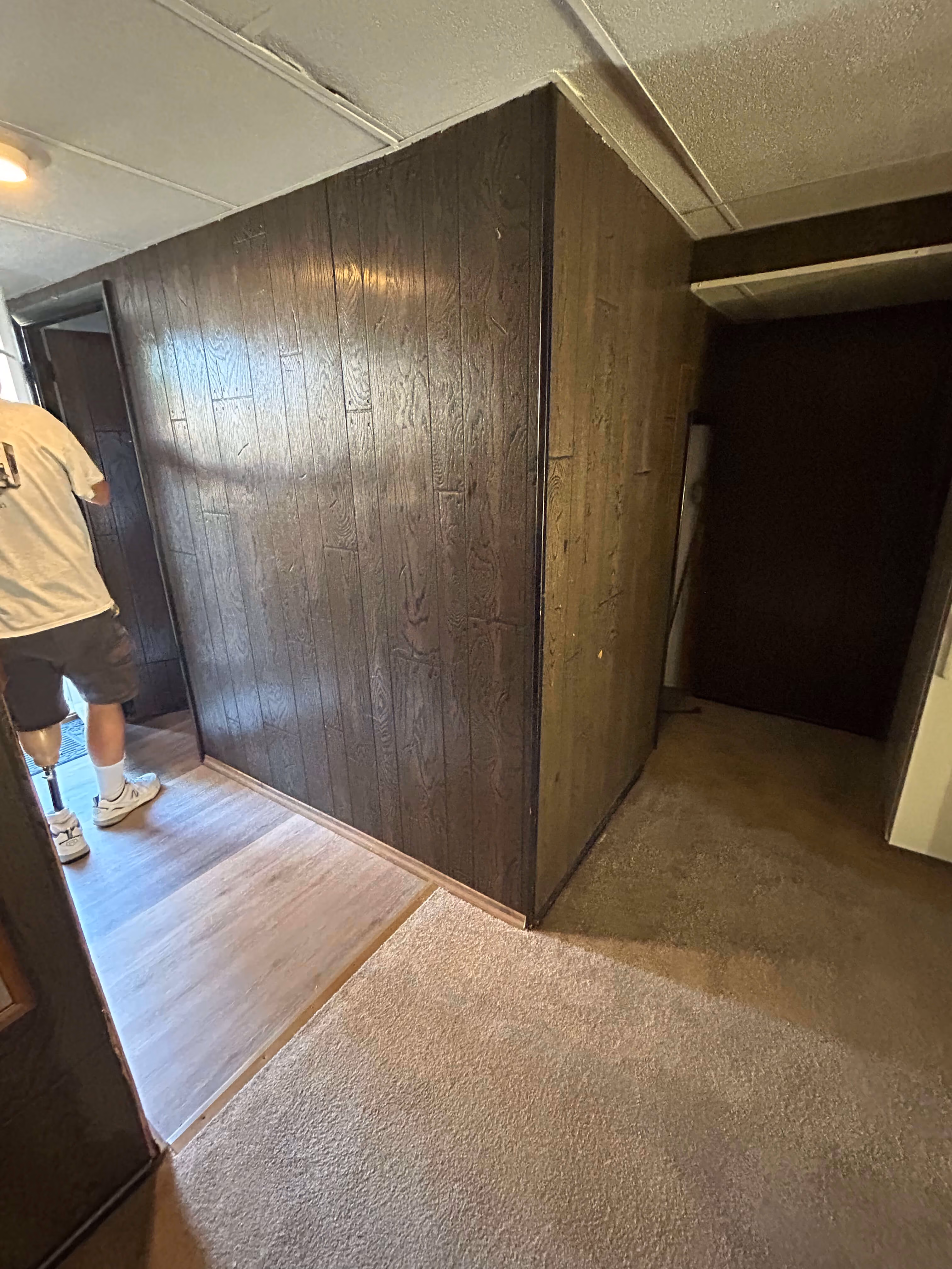 Interior corner with dark wood panel walls, carpet floor, and a person with a prosthetic leg.