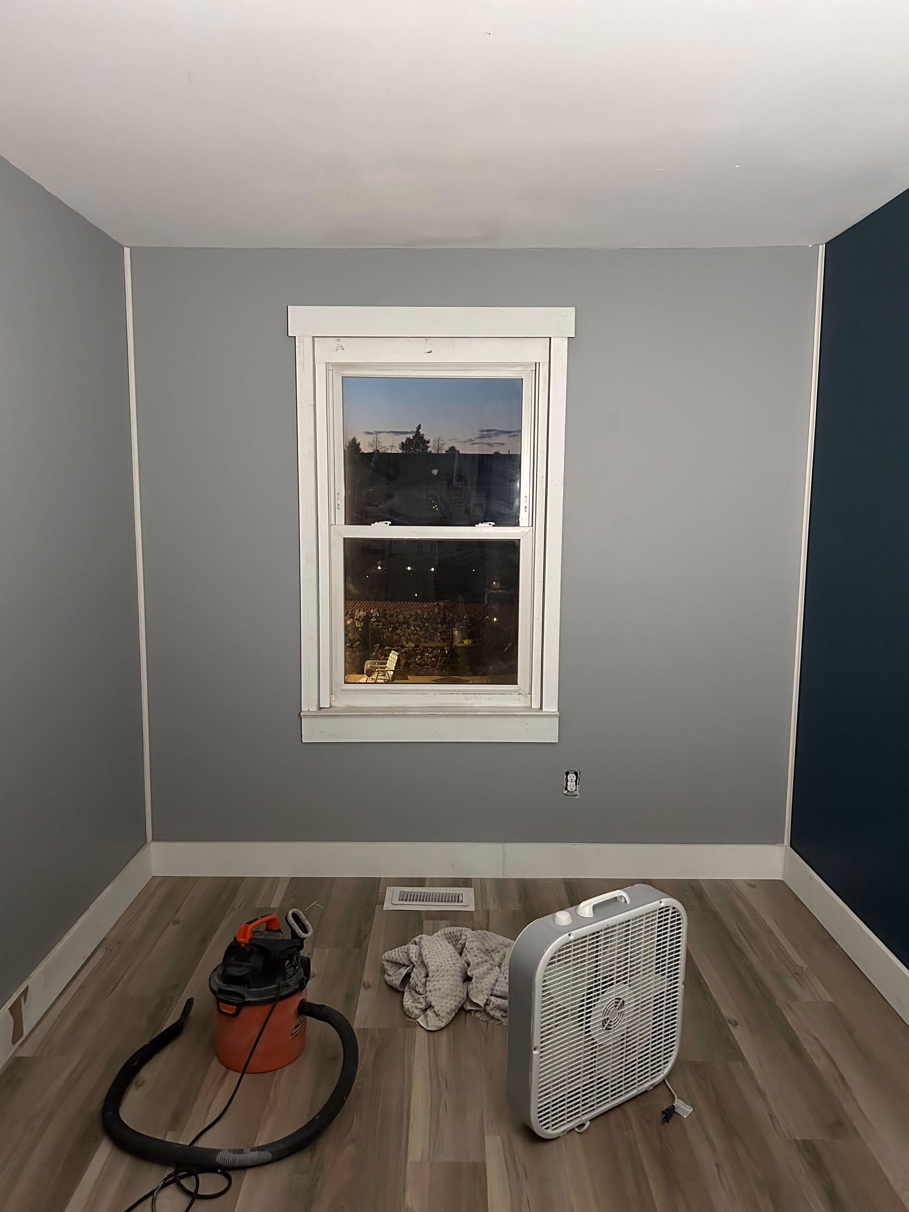 Empty room with gray walls, wooden floor, a window, a vacuum, a fan, and a cloth on the floor.
