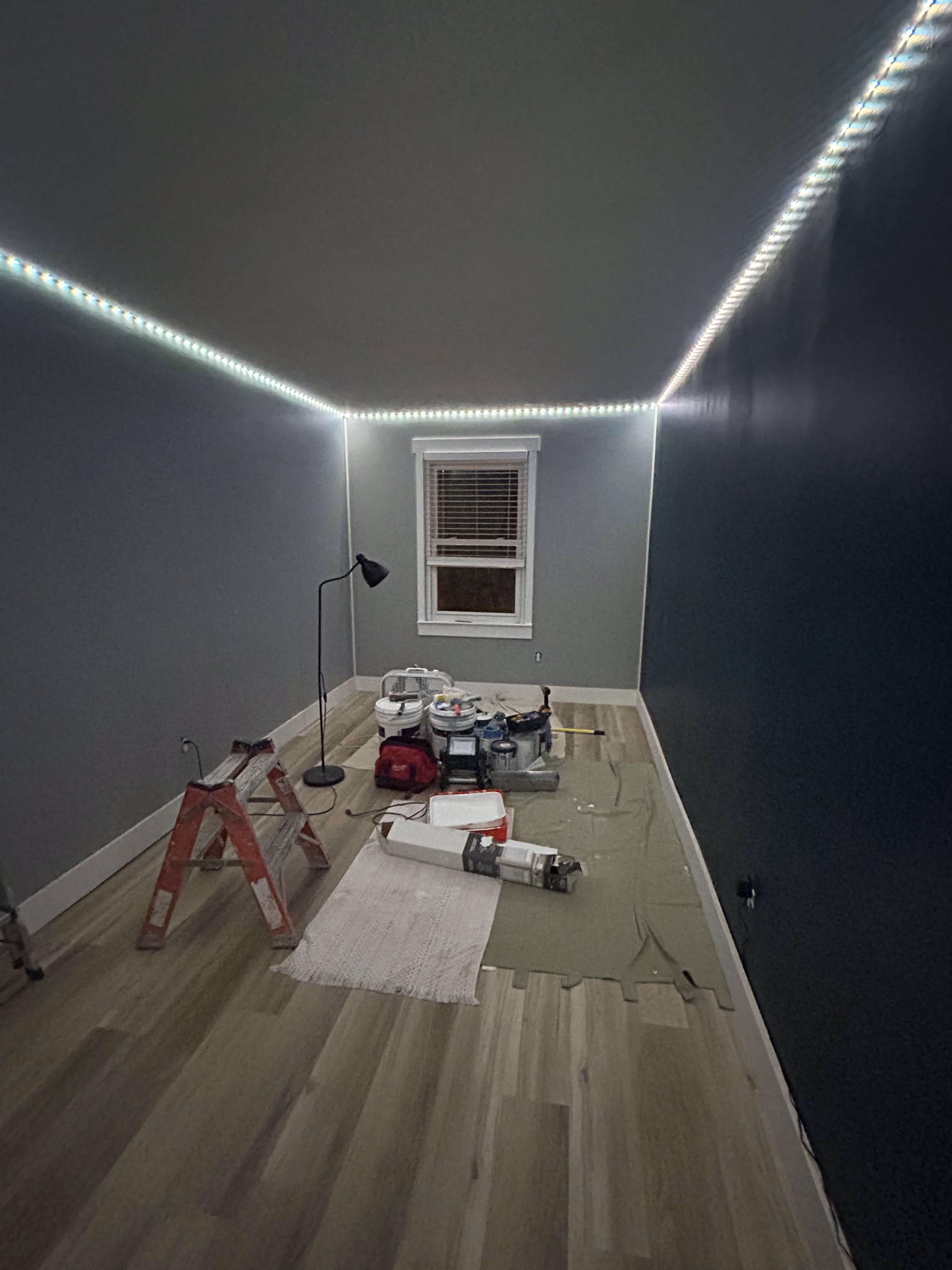 Room under renovation with gray walls, wooden floor, LED strip lights, ladder, and painting supplies.