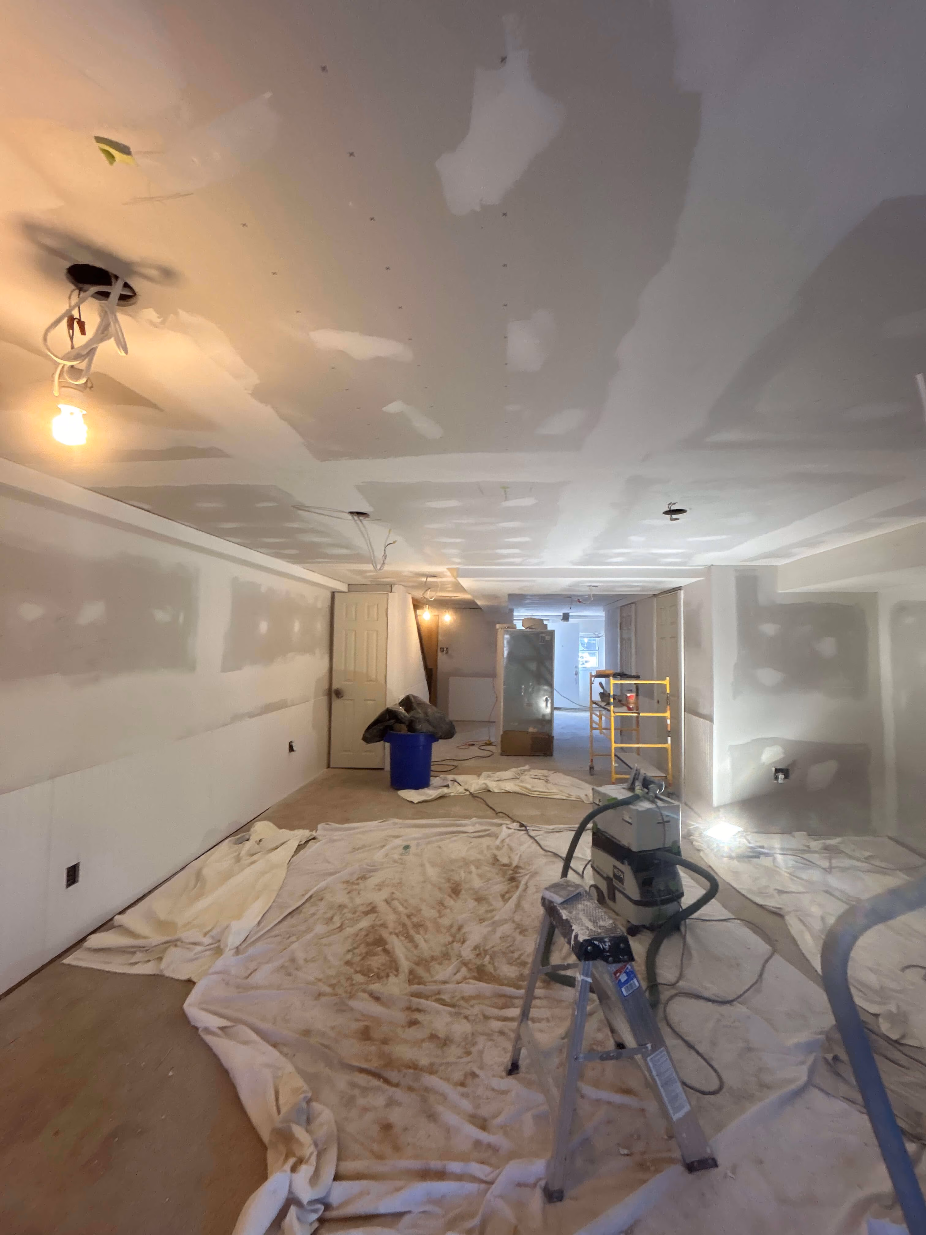 Room under construction with drywall installation, paint drop cloths, ladder, and construction equipment.