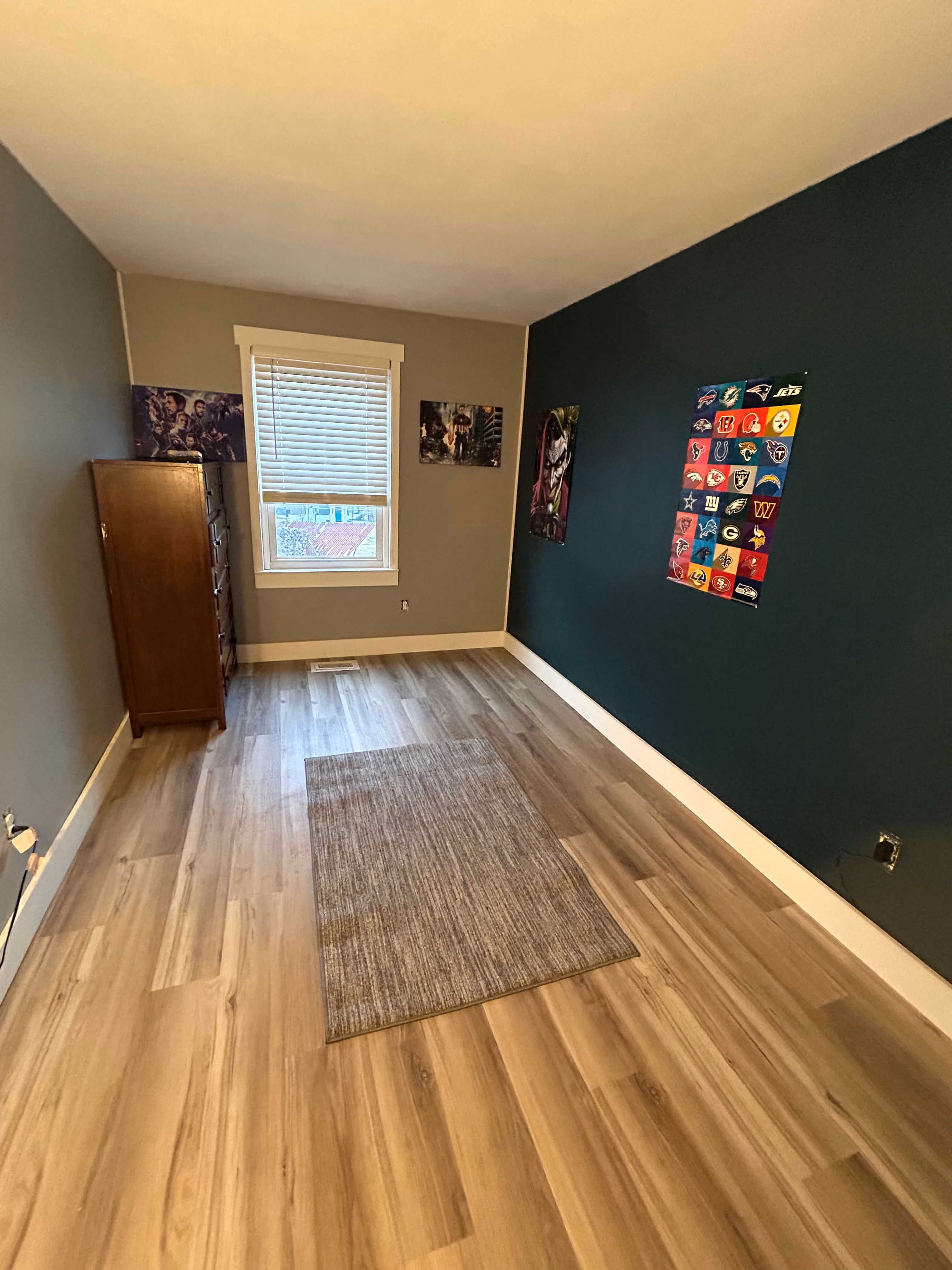 Empty room with wooden floor, small rug, brown dresser, window, and comic posters on walls.