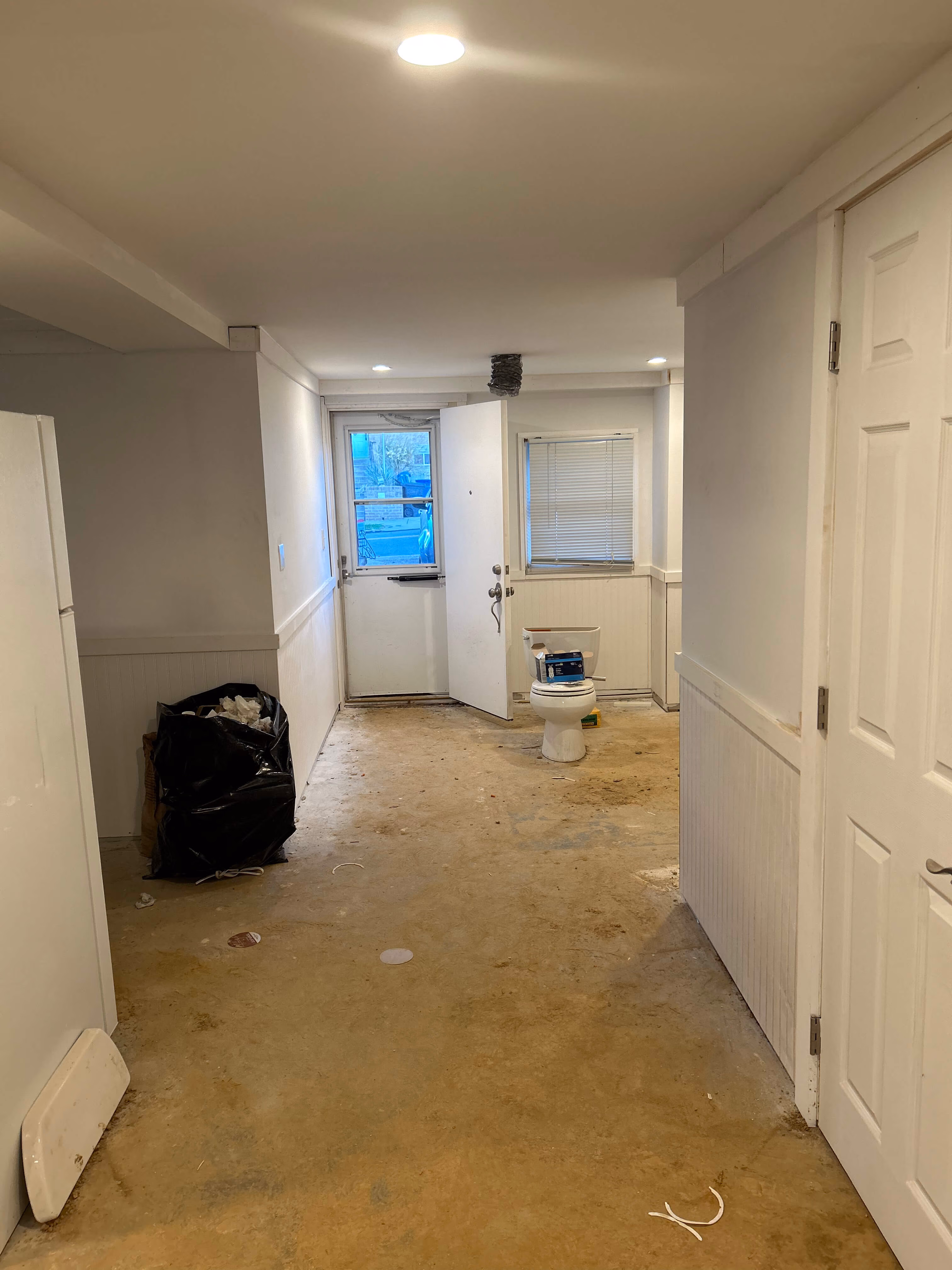 Empty room with a toilet, a trash bag, open door, and dirty floor under overhead lights.